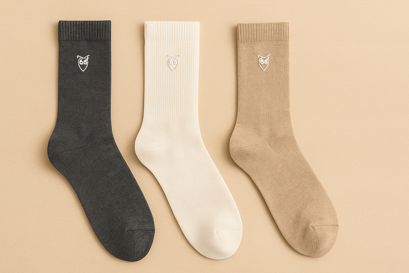 Drei Paar Knowledge Cotton Apparel Socken in den Farben Dunkelgrau, Off-White und Beige, nebeneinander auf einem neutralen, beigen Hintergrund – aus Bio-Baumwolle mit gesticktem Eulen-Logo.