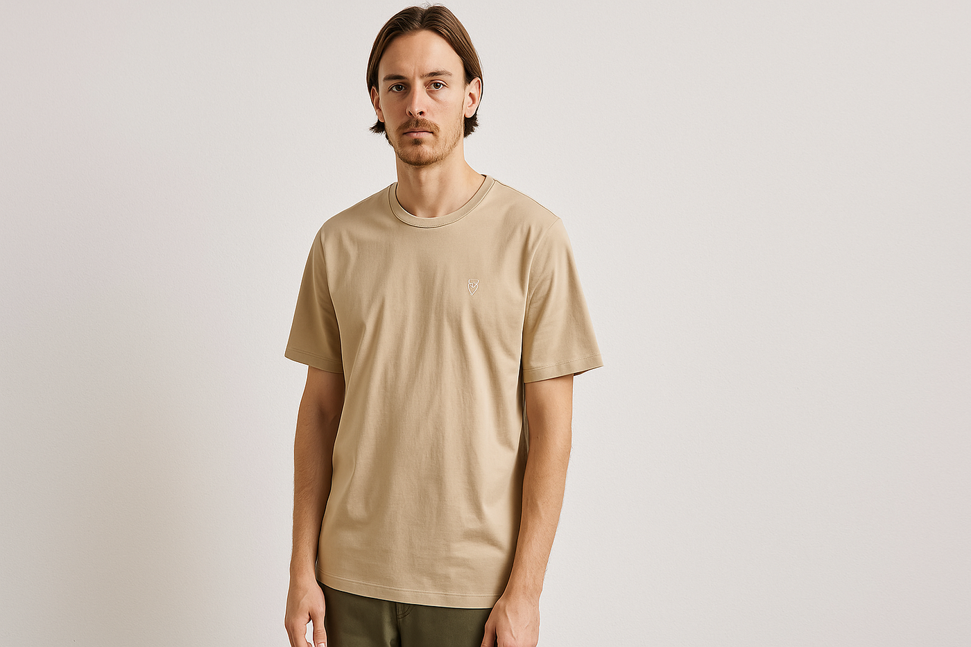 Junger Mann trägt ein beigefarbenes Bio-Baumwoll-T-Shirt von Knowledge Cotton Apparel mit gesticktem Eulen-Logo, kombiniert mit olivgrüner Hose, vor neutralem Hintergrund – minimalistischer, nachhaltiger Herrenlook.