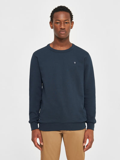 Mann trägt ein dunkelblaues Sweatshirt von Knowledge Cotton Apparel, Modell Erik Basic Badge in Total Eclipse, mit rundem Halsausschnitt und kleinem Brust-Logo.